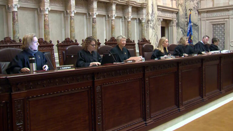 From left to right, Jill Karofsky, Rebecca Dallet, Ann Walsh Bradley Annette Ziegler, Rebecca Bradley, Brian Hagedorn and Janet Protasiewicz sit at a judicial dais in a row of high-backed leather chairs behind them, with another row of high-backed wood and leather chairs behind them, in a room with marble masonry and a Wisconsin flag.