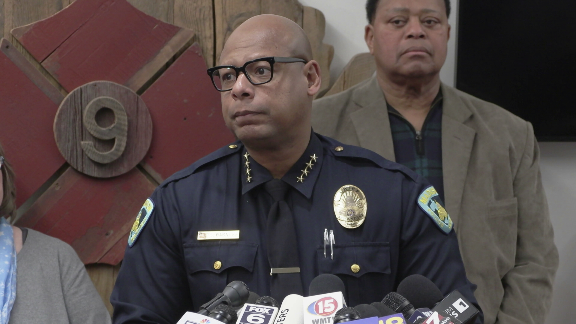 Shon Barnes speaks into multiple microphones with the flags of different media organizations while standing in a room with people standing next to and behind him and a wood fire department sign with the number "9" in the background.