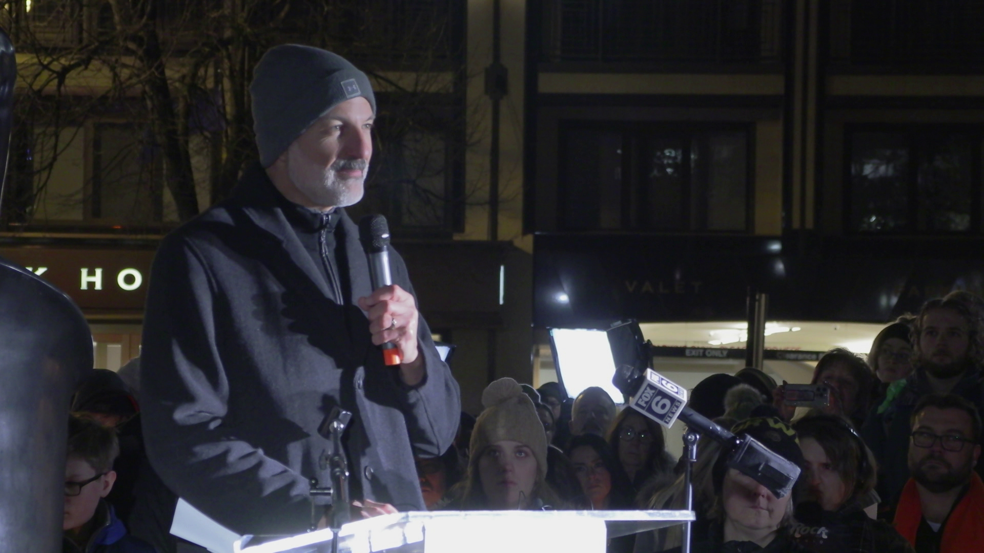 Joe Gothard speaks into a wireless microphone he holds with his left hand while standing outdoors behind a podium next to a group of people standing and a building in the background.