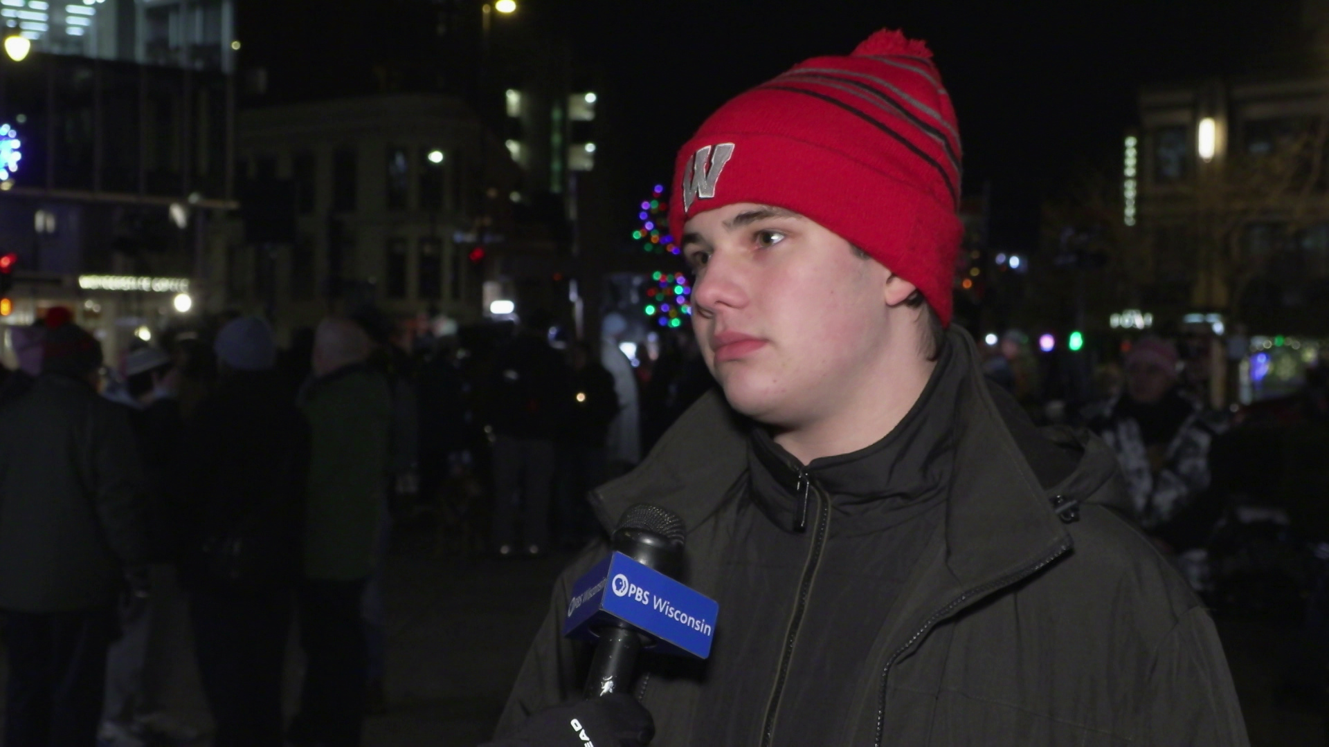 Jackson Ida speaks into a microphone with a "PBS Wisconsin" flag while standing outdoors with people, buildings and holiday lights in the background.