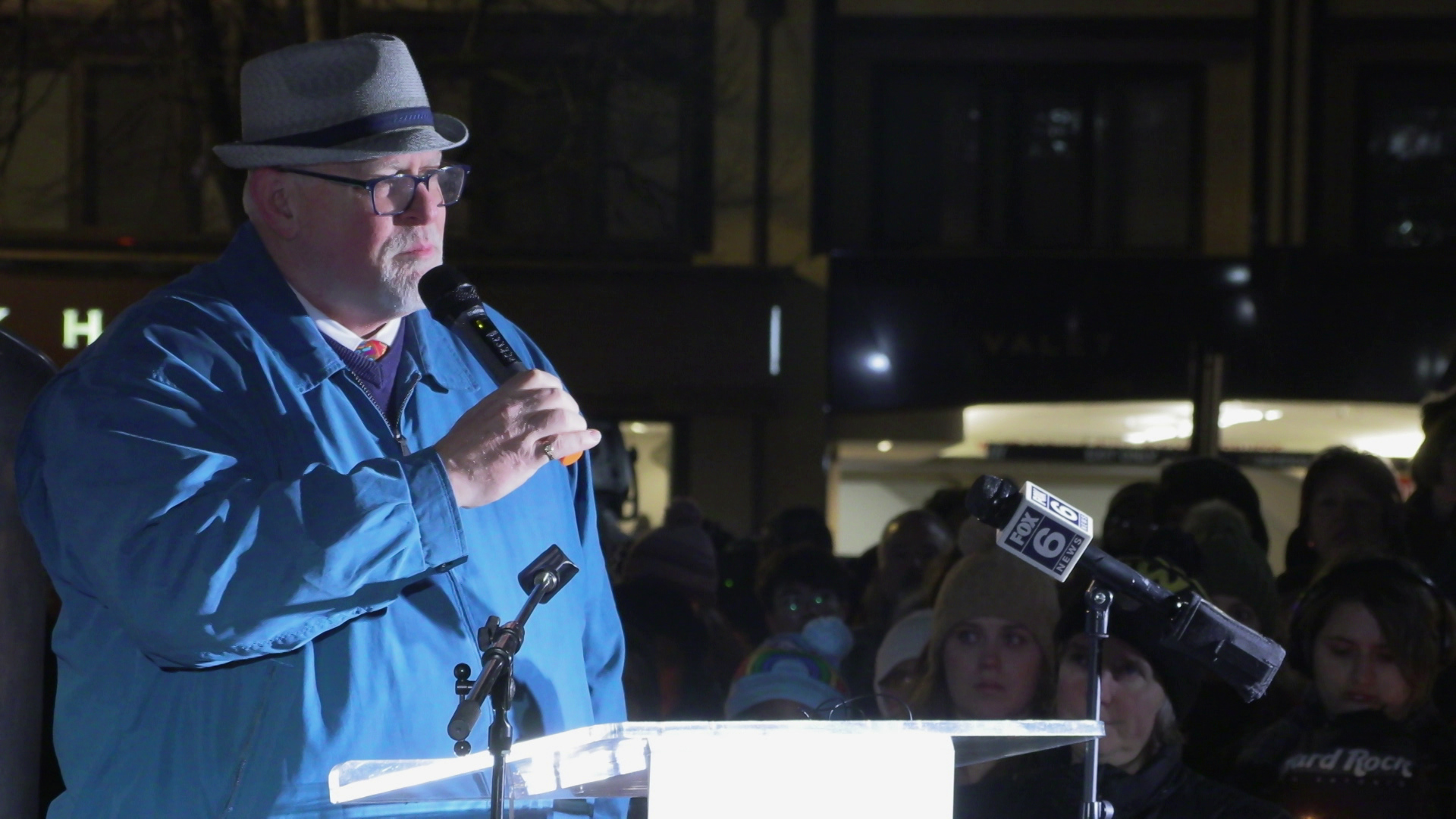 Chuck Moore speaks into a wireless microphone he holds with his right hand while standing outdoors behind a podium next to a group of people standing and a building in the background.