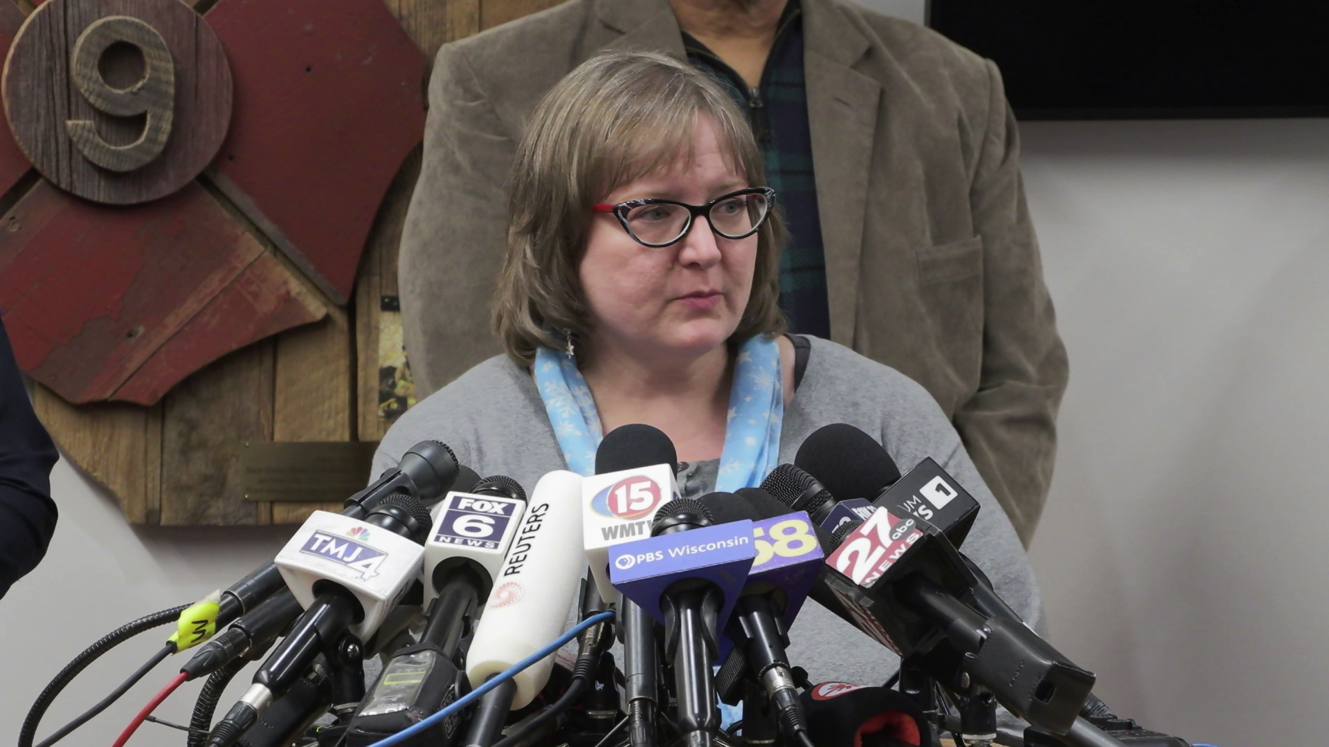 Barbara Wiers speaks into multiple microphones with the flags of different media organizations while standing in a room with people standing next to and behind him and a wood fire department sign with the number "9" in the background.