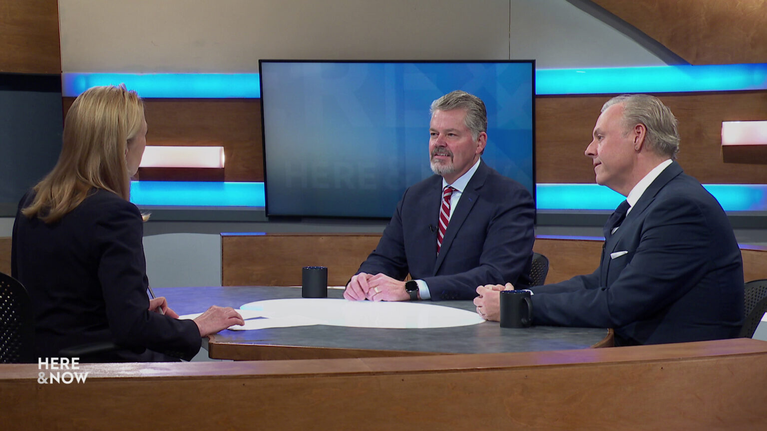 Bill McCoshen and Scot Ross sit next to each other and across from Frederica Freyberg at a desk on the Here & Now set.