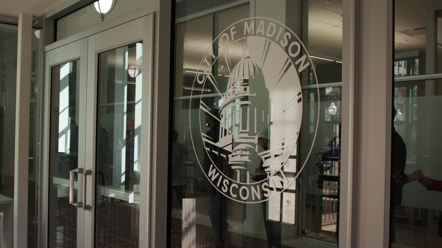 A window stencil of the City of Madison is attached to a large plate-glass window next to a pair of metal-framed double doors, people standing in line visible inside.