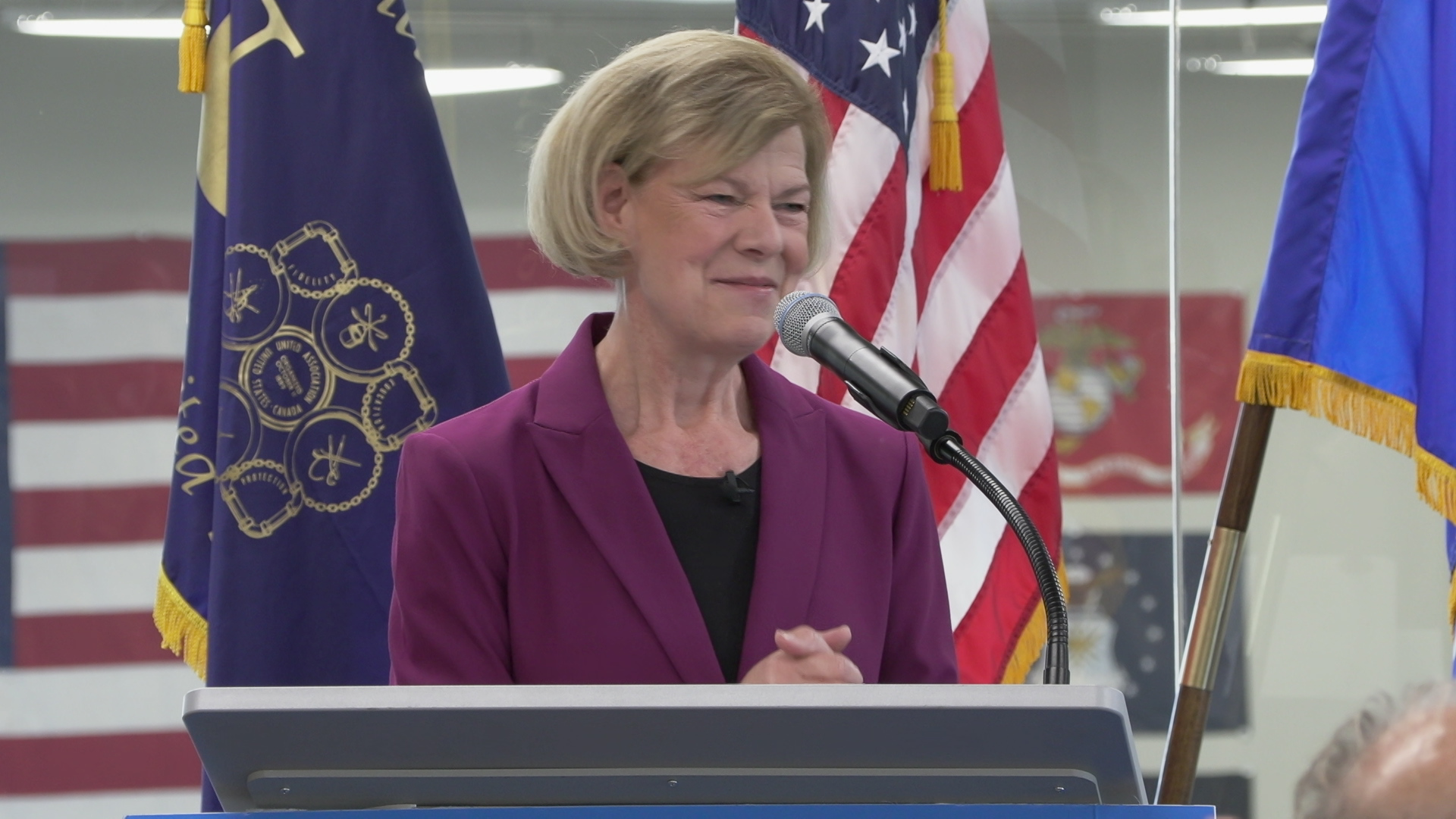 Tammy Baldwin speaks into a microphone mounted to a podium, while standing in a room with glass panels and flags in the background.