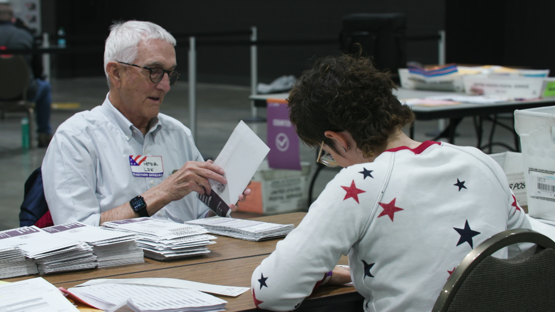 Two people hold absentee ballot envelopes while seated at a table facing each other, with tables and boxes of ballots and papers in the background in a room with a concrete floor.