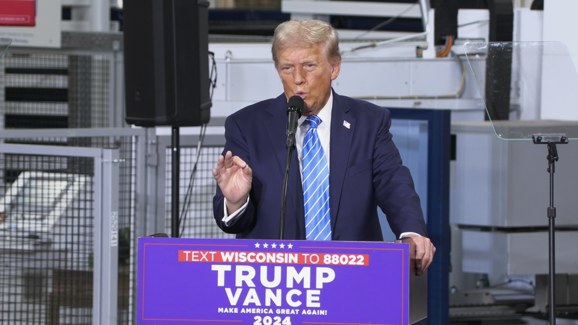 Donald Trump gestures with his right hand while speaking into a microphone mounted to a podium with a sign reading "Trump Vance," while standing and facing a teleprompter mirror, in a room with a speaker and industrial machinery in the background.