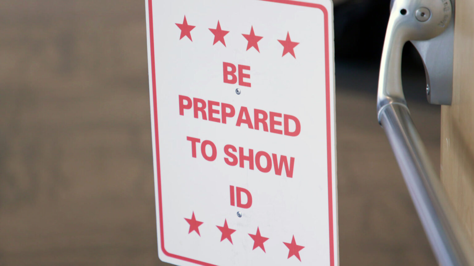 A metal sign with the words Be Prepared to Show ID framed above and below by lines of four stars stands next to a swinging metal push and pull commercial door handle.