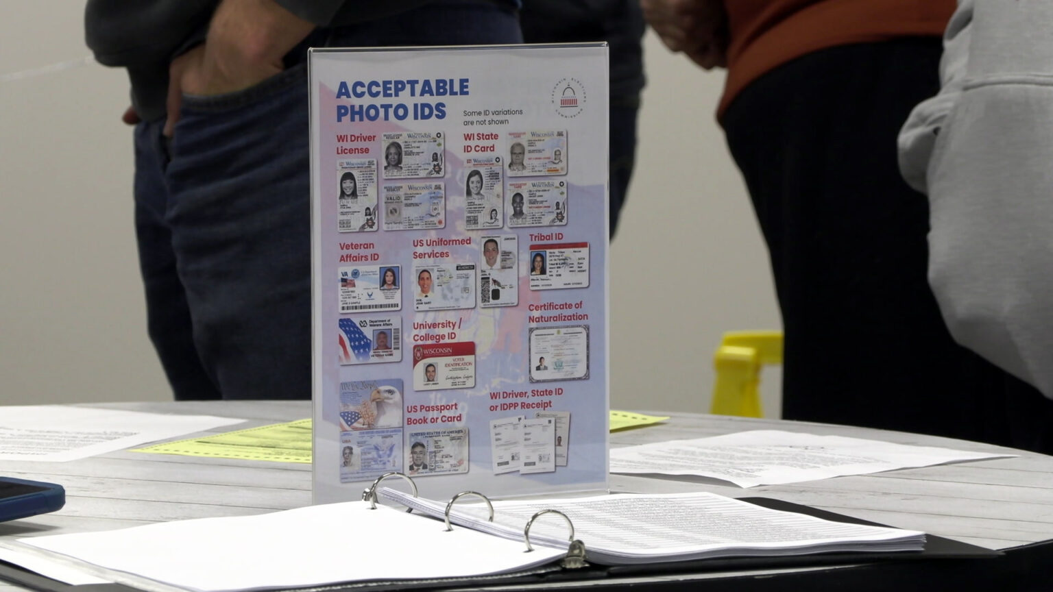 A piece of paper with the title Acceptable Photo IDs and graphics of different forms of photo identification that is placed in a transparent display stands at the center of a table with papers and a 3-ring binder on its surface, with  people standing in the background.