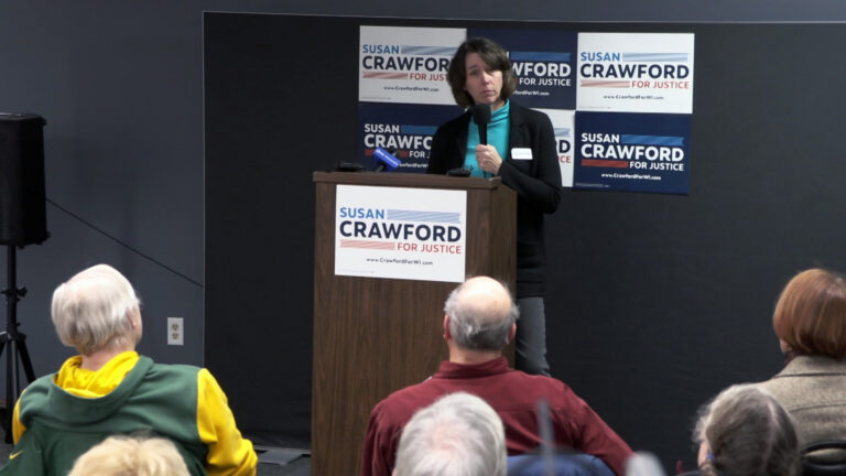 Susan Crawford holds a microphone in her right hand and speaks while standing behind a wood podium with other microphones on its surface and a campaign sign reading Susan Crawford for Justice on its front, with six more campaign signs displayed on a wall behind her, with a speaker on a stand to one side and people seated and facing her in the foreground.