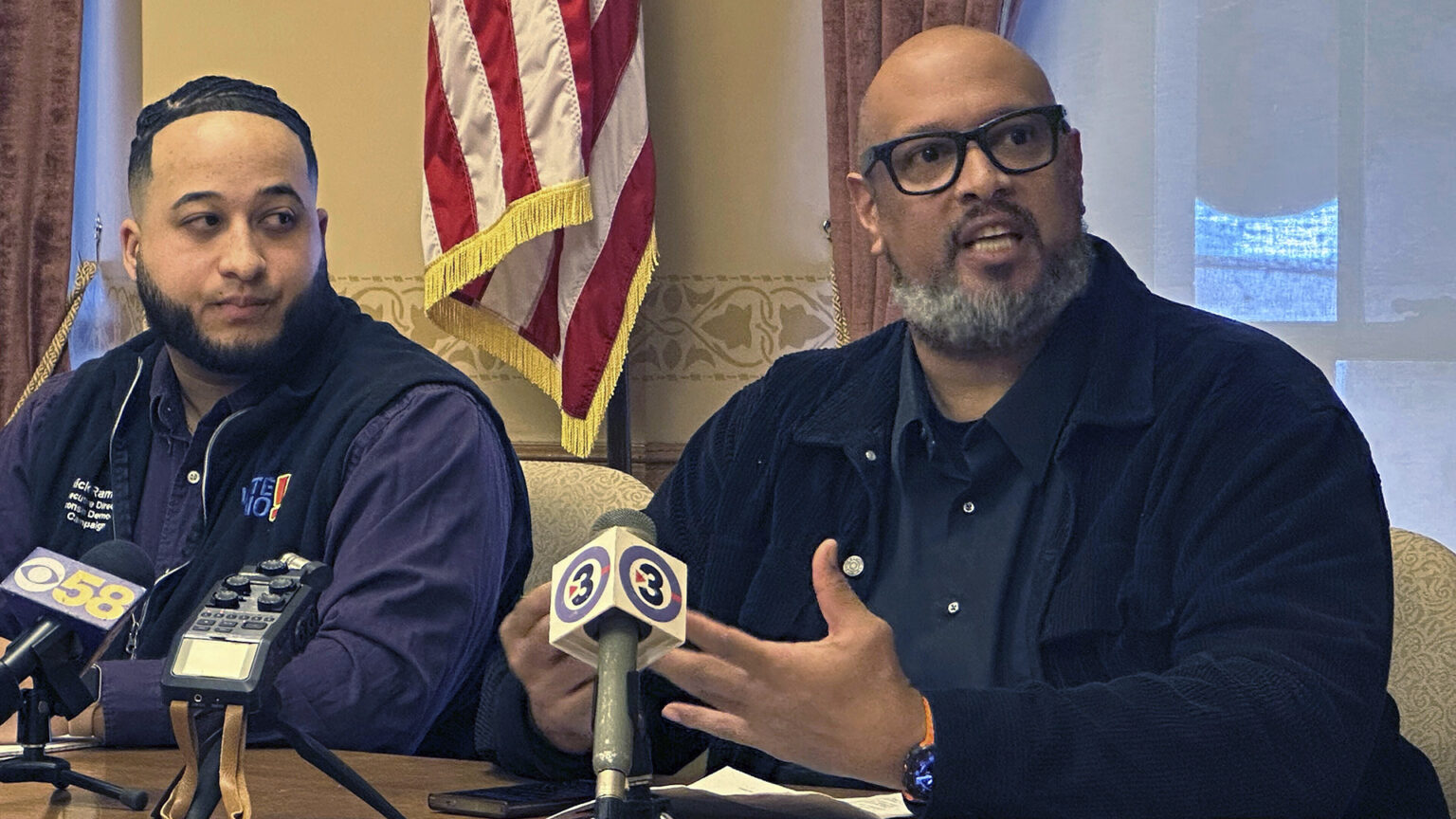 Harry Dunn gestures with both hands while sitting at a table with multiple microphones with flags of different media organizations on its surface, with another person seated to the side in a room with windows with open drapes and a U.S. flag in the background.