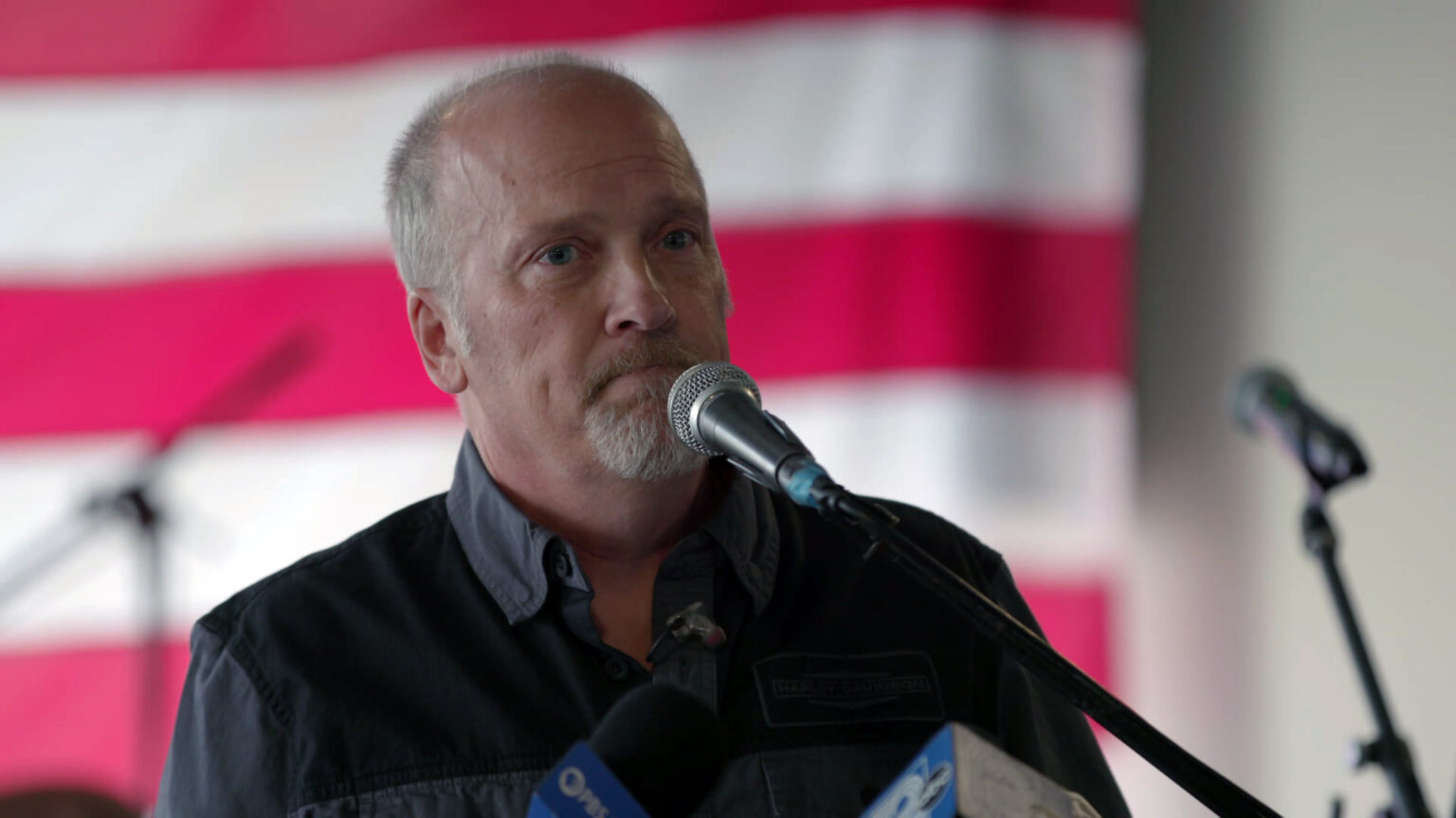 Brad Schimel stands and speaks into multiple microphones, with out-of-focus microphones, microphone stands and the strips of a U.S. flag in the background.