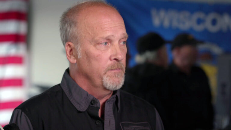 Brad Schimel stands and speaks with out-of-focus people standing in the background of a room with the U.S. and Wisconsin flags displayed flat on a wall.