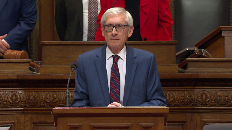 Tony Evers stands with this hands crossed on the top of a wood podium with a microphone mounted to its surface, with three other people standing behind him on another tier of a wood legislative dais with inlay carvings.