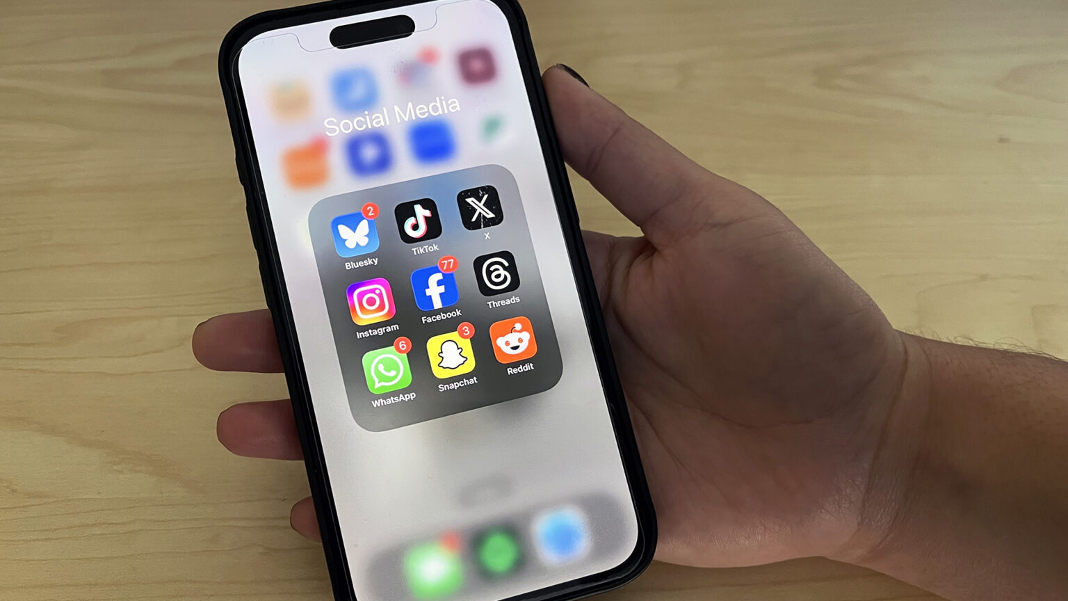 A hand resting on a blonde wood tabletop holds a smartphone showing a screen with a menu of the social media apps Bluesky, TikTok, X, Instagram, Facebook, Threads, WhatsApp, Snapchat and Reddit.