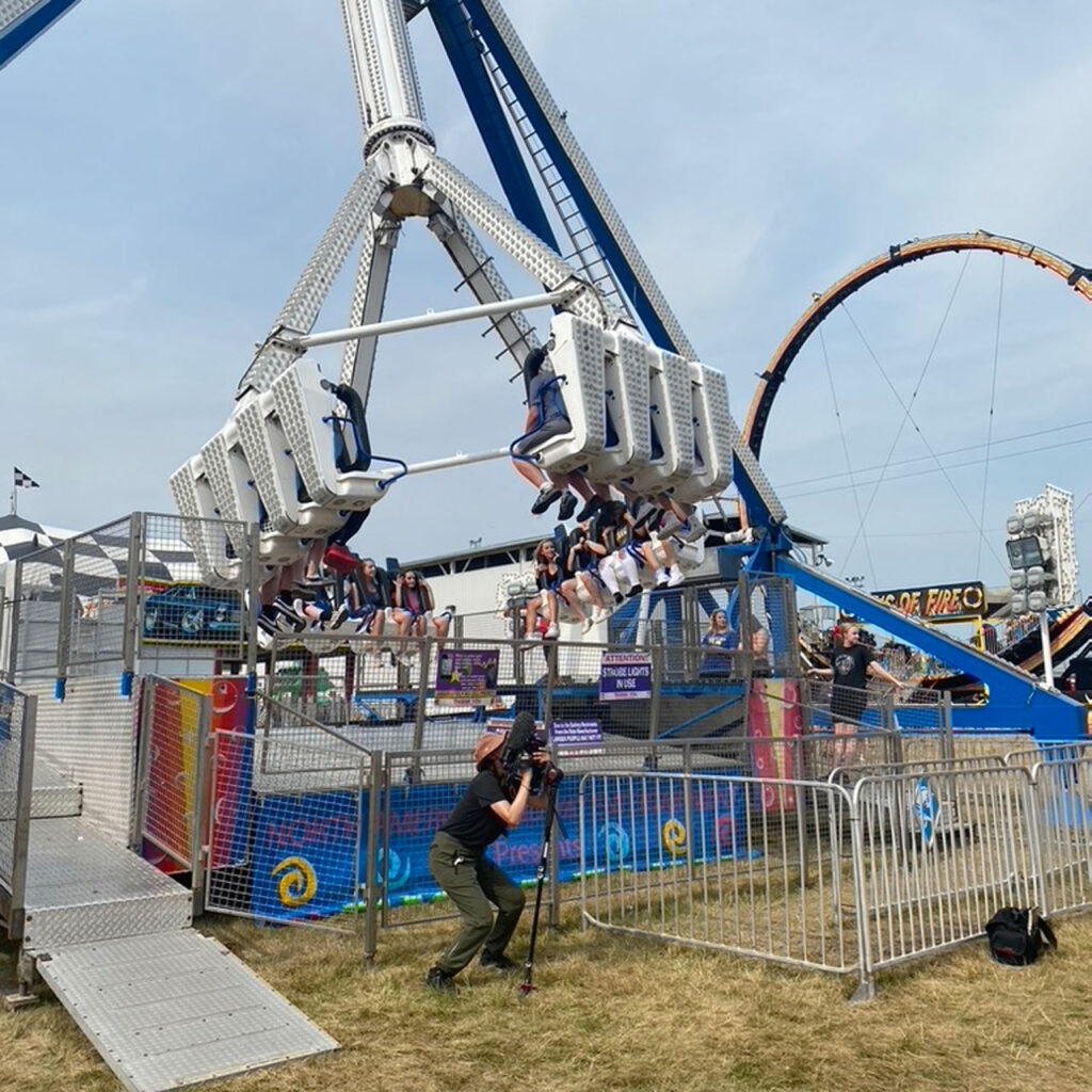 A woman stands with a tripod and records close footage of amusement rides at a fair.