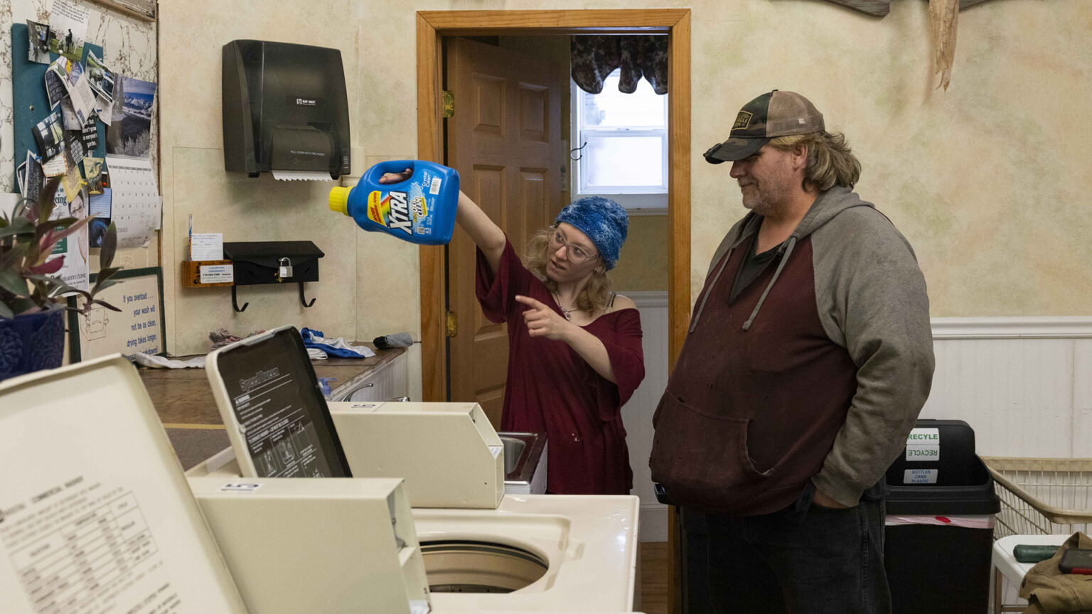 Christina Hubbell holds a plastic jug of laundry detergent up in the air while standing next to Eric Zieroth inside a room with a washing machine and dryer, a plastic recycling bin, a sink and a wall-mounted paper towel dispenser next to a bulletin board with a calendar and other items pinned to its face, with an open wood door showing a window in an adjacent room.