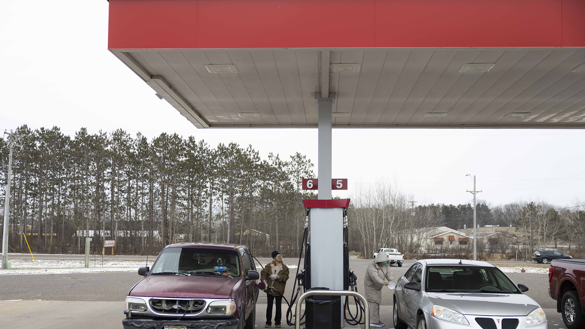 Christina Hubbel stands pumps fuel while standing next to a car parked at a gas pump, with another motorist on the other side, with a parking lot, road, trees and buildings in the background.