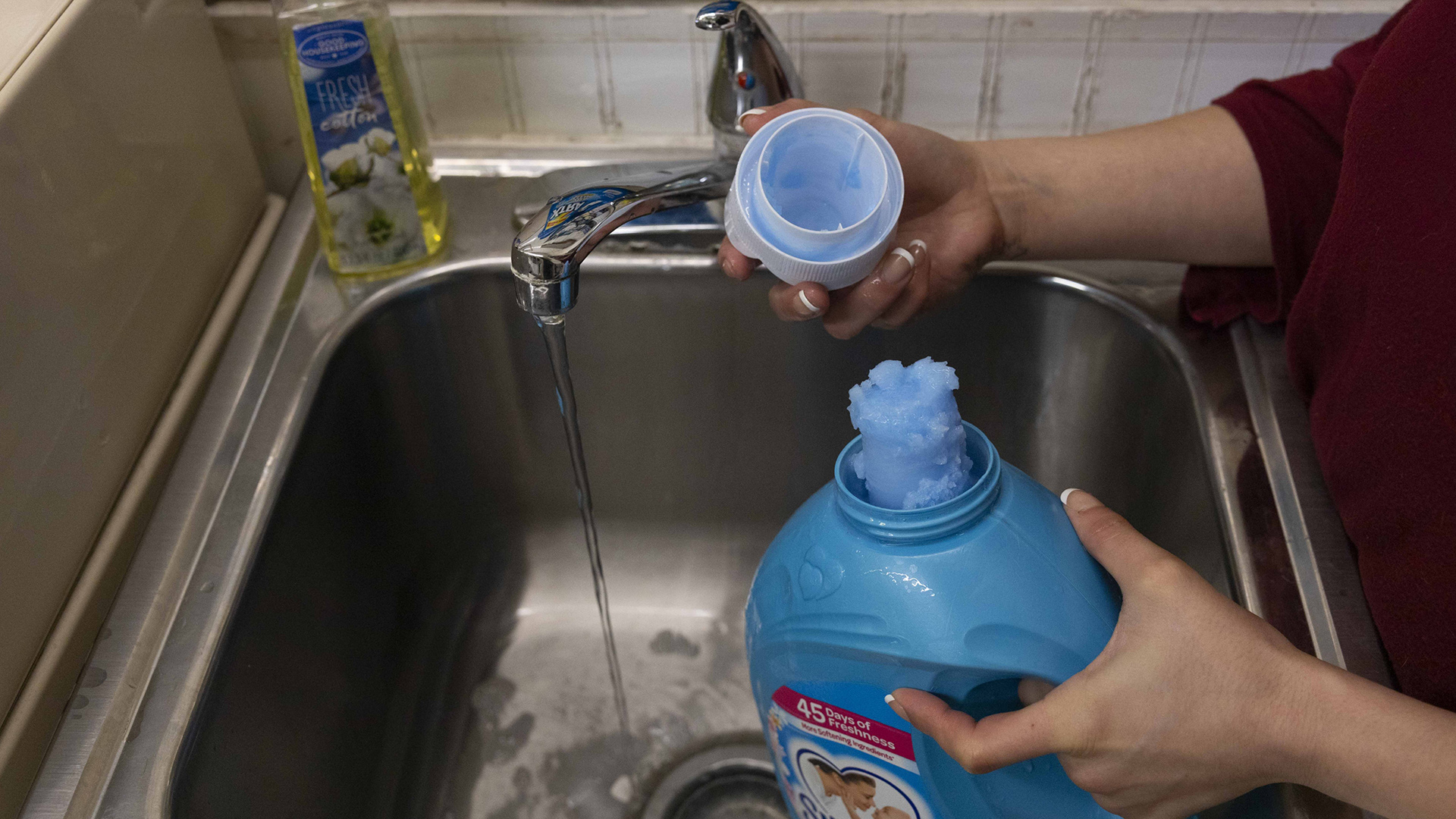 Christina Hubbell holds a plastic laundry detergent container with a column of frozen detergent at its top in her left hand and the container's cap in her right hand, standing over a stainless steel washbasin with a stream of water flowing from a faucet.