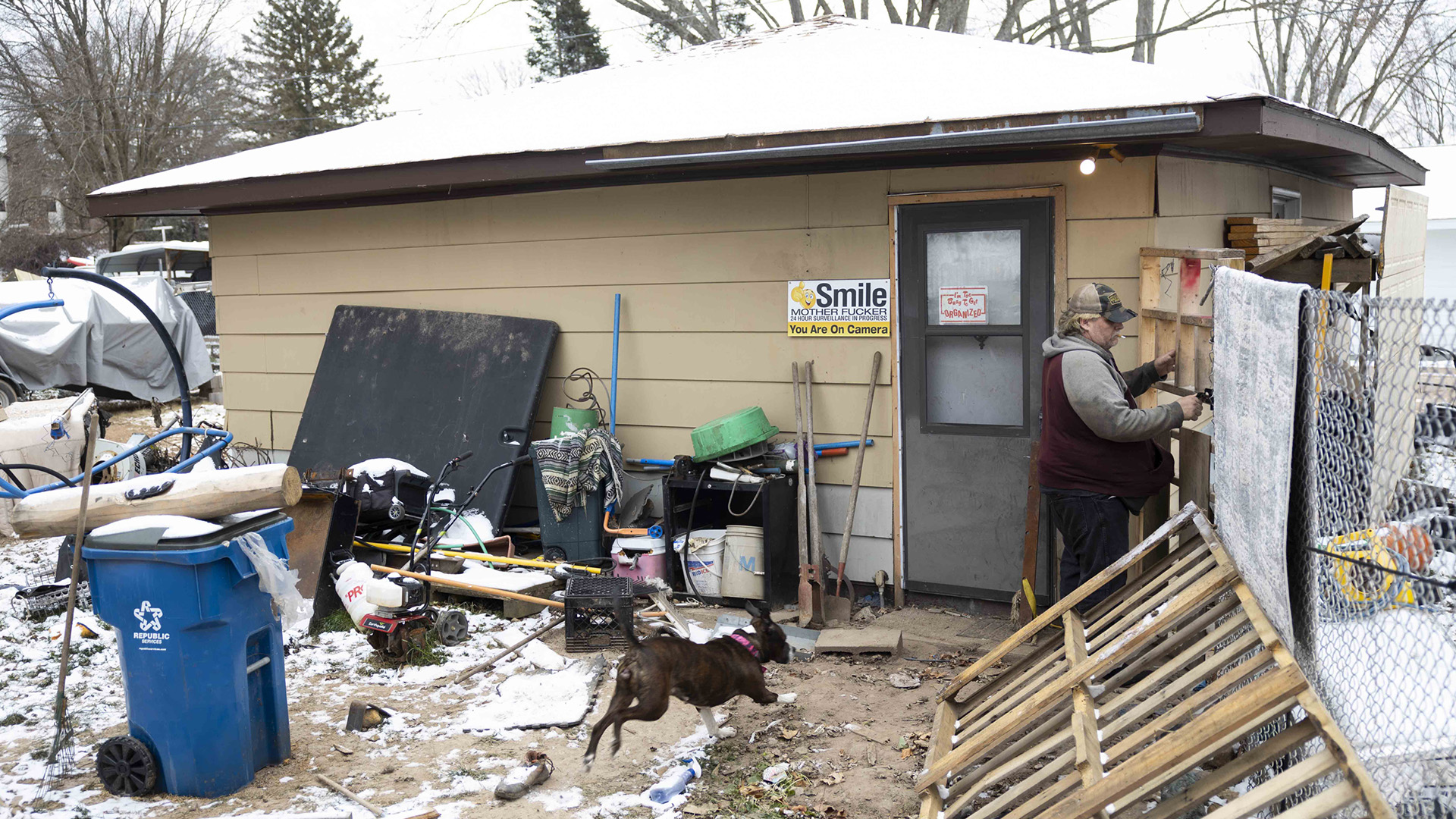 Eric Zieroth stands next to a wood gate attached to the corner of a small single-story building with a door on one side and a chain link fence on the other side, with a dog, recycling bin with wheels, wood pallets and other items in the yard in front of the building, and with parked vehicles and trees in the background.