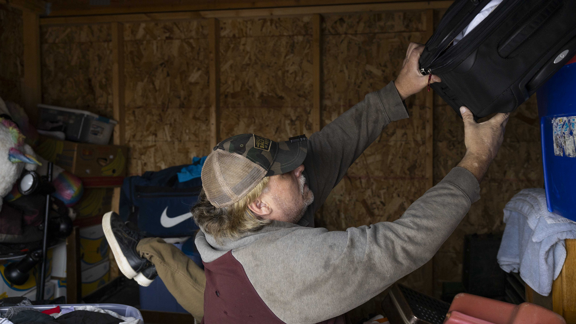 Eric Zieroth pulls a suitcase down from a stack of multiple items, with other stacked items in the background of a room with plywood walls.