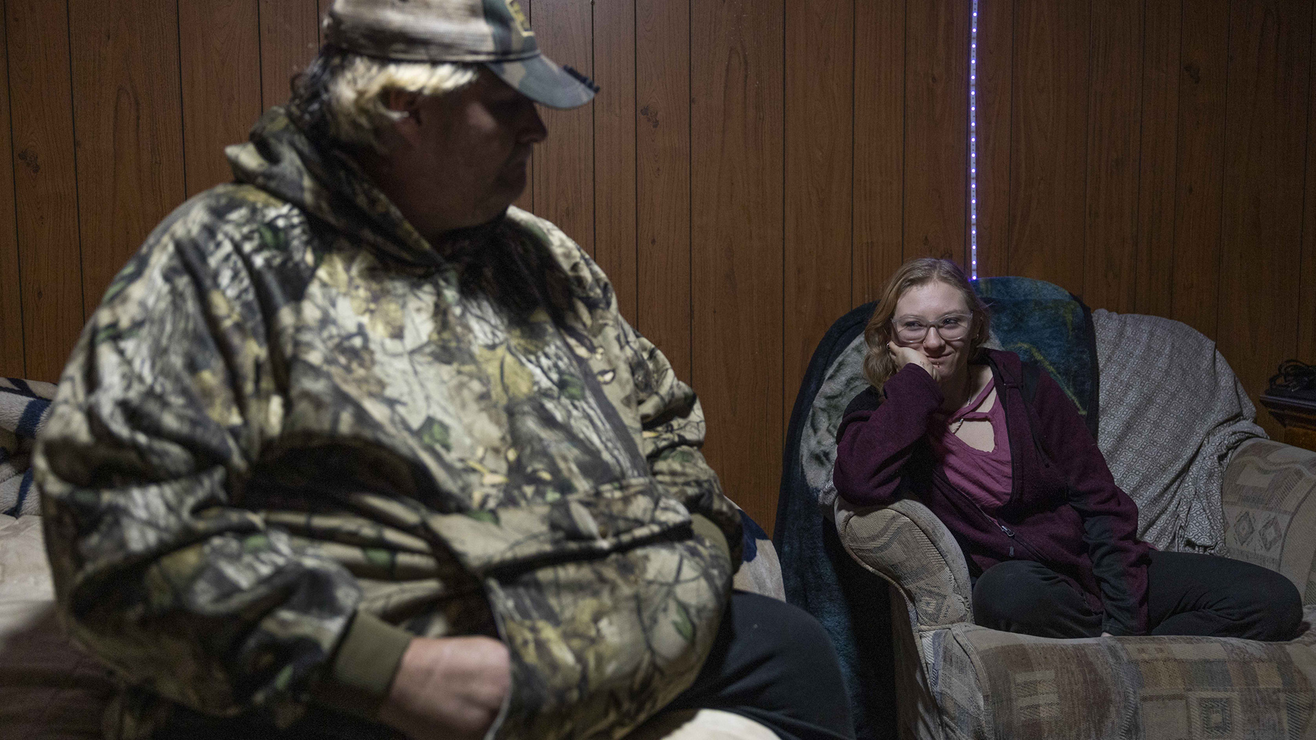 Christine Hubbell sits atop a cloth-upholstered chair with sidearms and rests her chin in her right hand while an out-of-focus Eric Zieroth sits in the foreground, in a room with laminate wood-paneled walls and a LED light string.