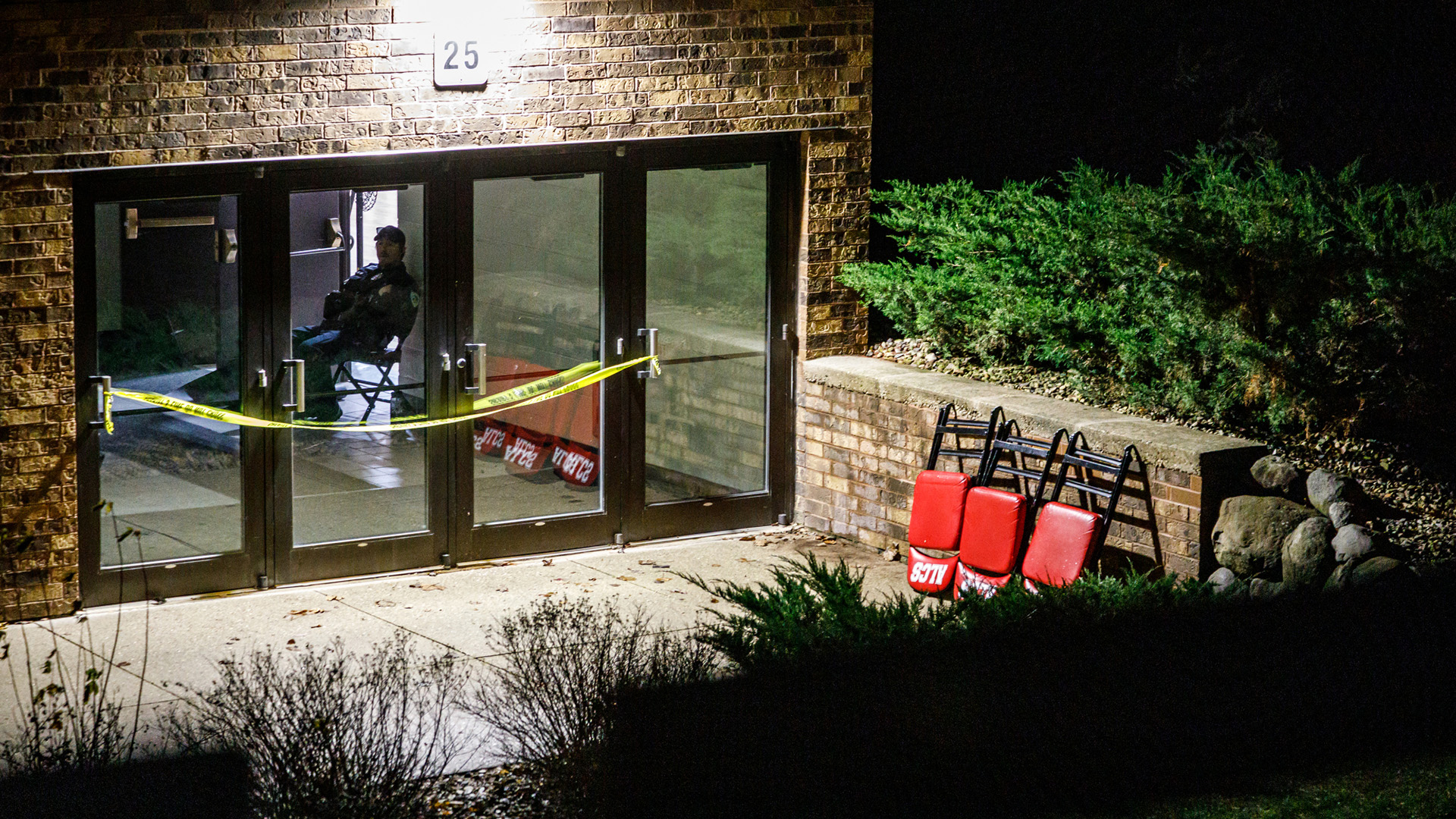 Police tape is strung across the handles of four metal-framed glass doors at an entrance to a brick building with folded chairs set against a brick retaining wall next to an exterior concrete patio with landscaping plants to the side and in the foreground illuminated by a building light, with a police officer seated on a folding chair inside the building next to two other doors that have been propped open.