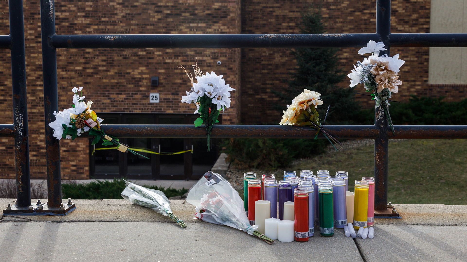 A spontaneous memorial features multiple flower bouquets attached to vertical and horizontal metal poles that comprise a railing, and more flower bouquets, multiple devotional candles and flameless candles placed on a concrete sidewalk next to a lawn, with a lawn and brick building in the background.