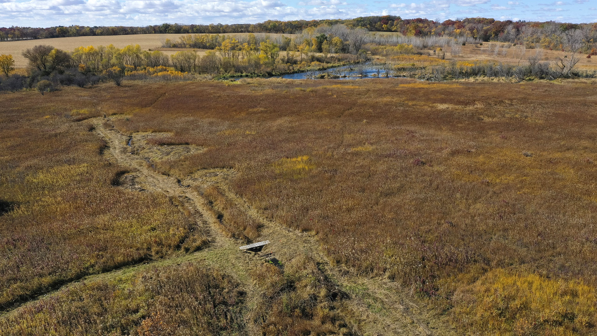 An aerial photo shows a line of disturbed ground with multiple circular spurs in the midst a flat area of prairie and marsh, with a waterway with trees along its banks, a field and wooded areas in the background under a mostly cloudy sky.