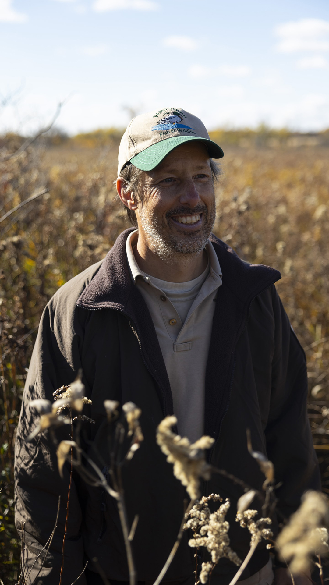 Mike Engel smiles walk walking amid foliage in a field under an out-of-focus partly cloudy sky.