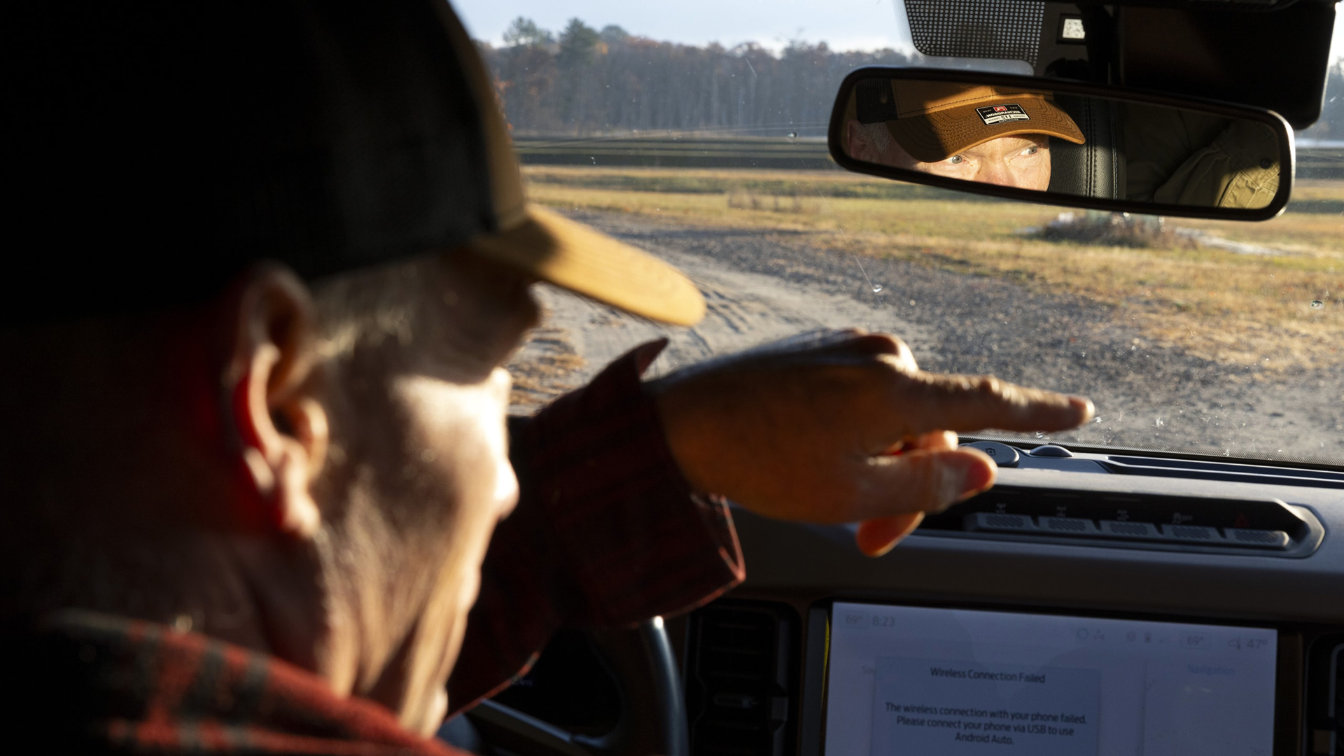 Jim Hoffman sits at the wheel of a vehicle and points with the index finger of his left hand above a dashboard touchscreen display, with his eyes and nose visible in a rearview mirror mounted in a front windshield, through which a dirt road and line of trees on the horizon are visible.