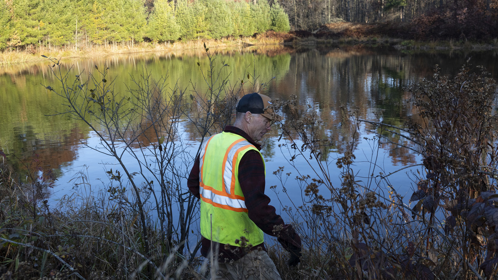 Jim Hoffman wears a fluorescent safety vest while standing amid foliage at the end of a pond with calm water, with conifer trees along its opposite shore in the background.