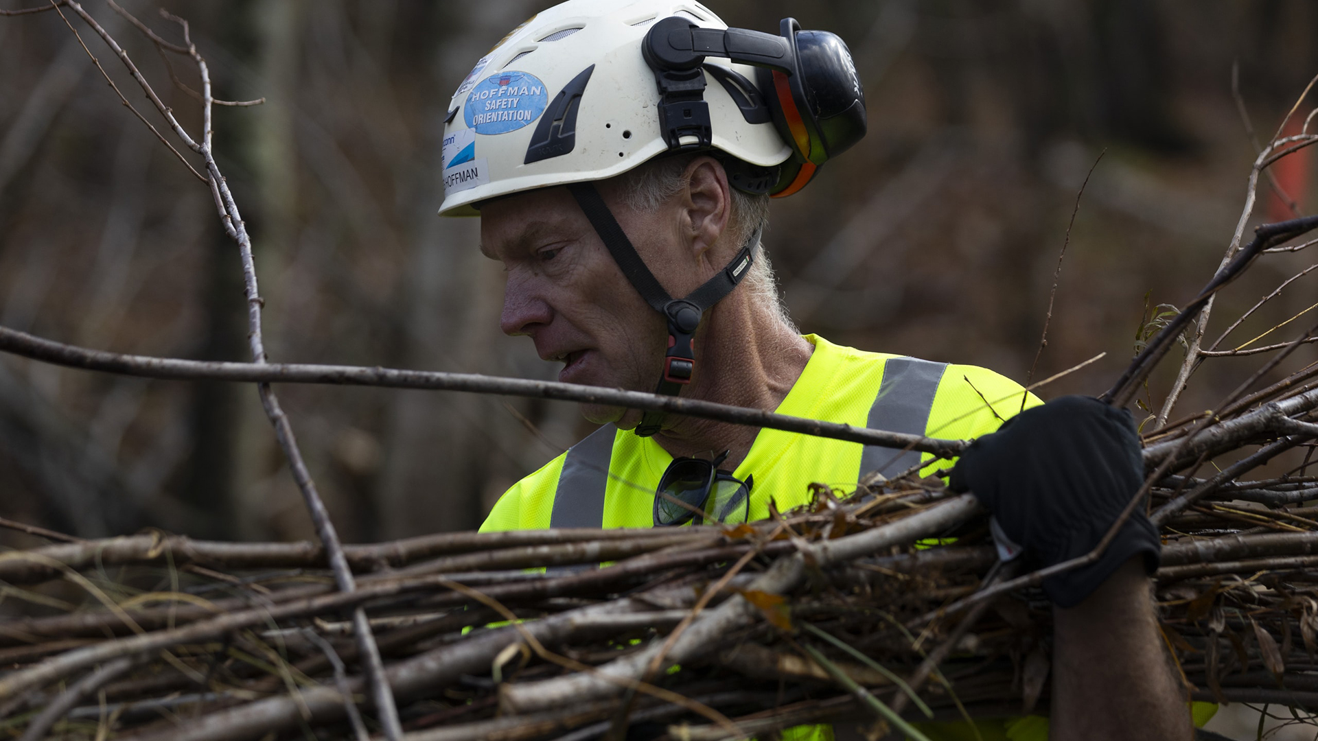 Jim Hoffman carries holds a bundle of sticks in his gloved left hand and arm while wearing a safety helmet with a chinstrap and rear-mounted light.