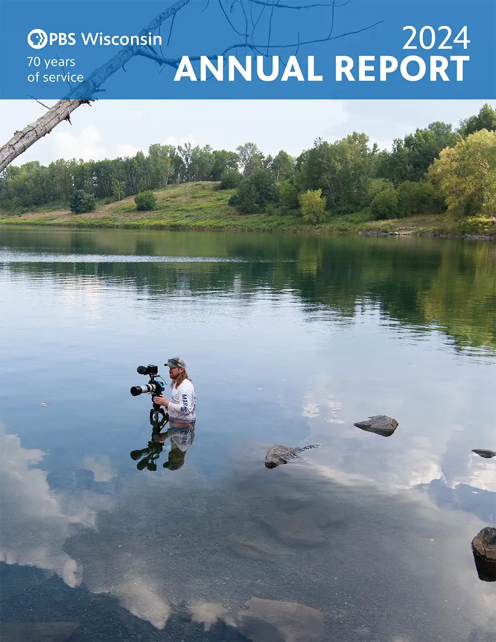 A photographer stands waist-deep in a calm lake, filming with a professional video camera. The image features a "PBS Wisconsin 2024 Annual Report" banner.