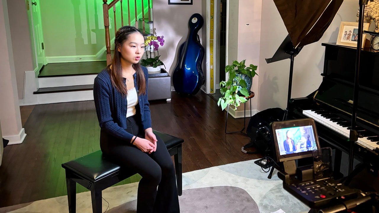 A teenage girl sitting at a piano bench in her home, being interviewed by a camera crew.