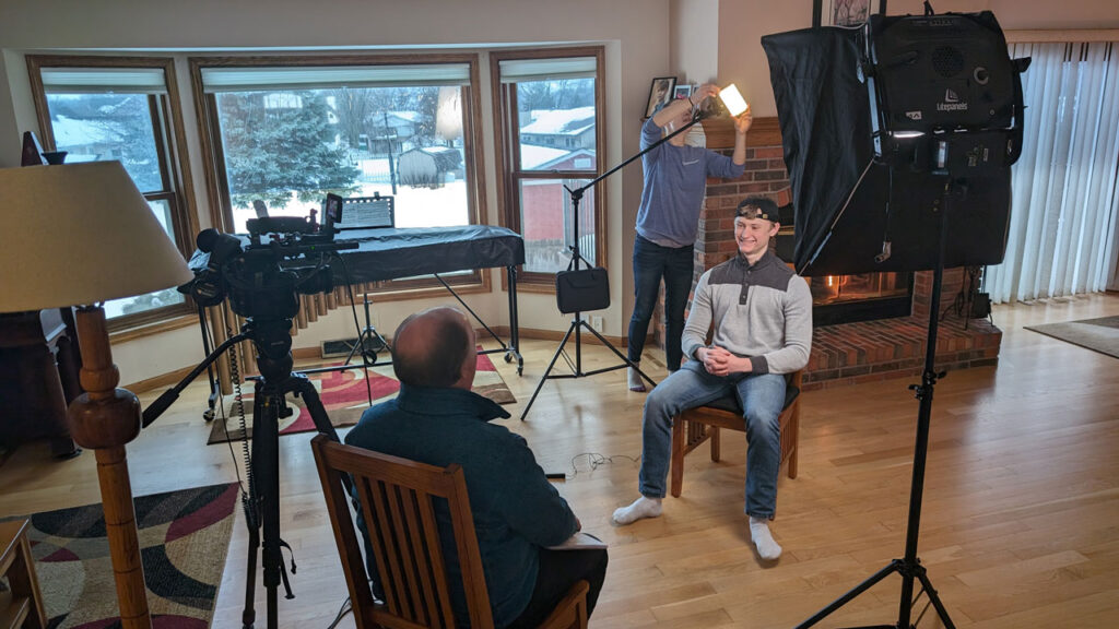 A teenage boy is interviewed in his living room by a man sitting in a chair while a woman sets up studio lighting.