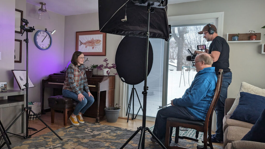 A teenage girl is interviewed sitting at a piano in her home by two men setting up camera equipment.