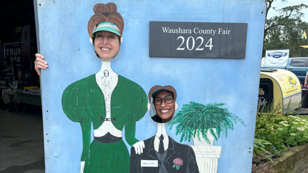 Two women smile as they pose in a cartoonish wooden cut-out photo-op of two people in period costume.