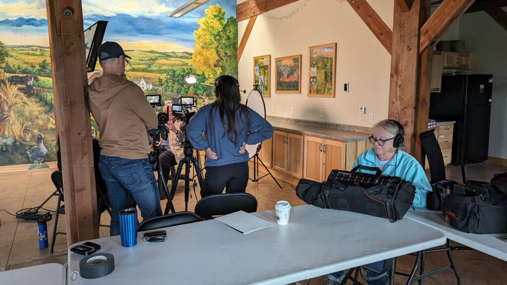 A production team monitors a video camera and sound equipment during an interview inside a rustic conference room.