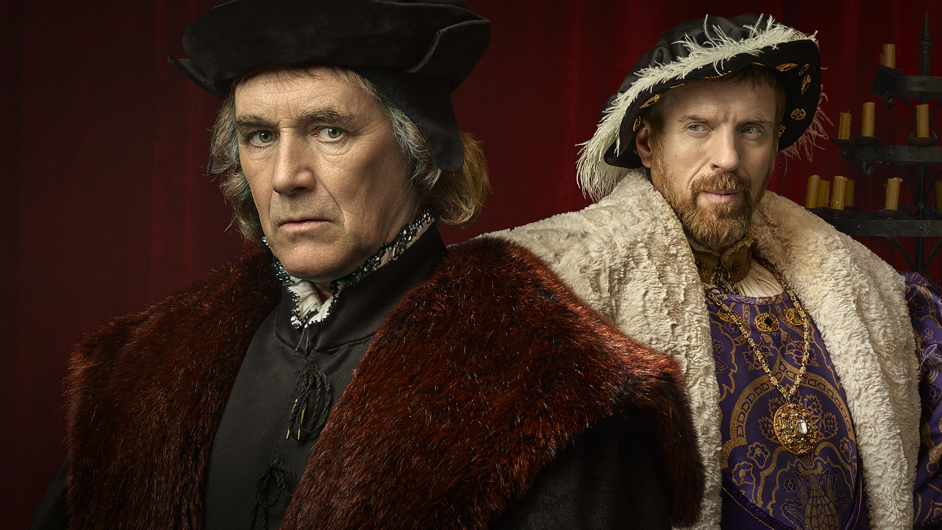 A promotional image from *Wolf Hall: The Mirror and the Light* featuring two men in elaborate Tudor-era clothing. The man in the foreground wears a dark fur-lined coat and a black cap, with a serious expression. The man in the background, dressed in regal purple and gold with a fur cape and feathered hat, looks confidently ahead. The setting is dimly lit with a red curtain and candles in the background, evoking a historical atmosphere.