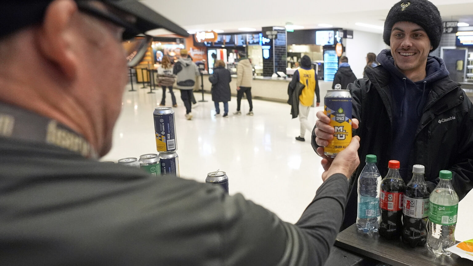 Jimmy Gross hands a tall-boy beer can with the logos of Third Space Brewing and Marquette University on its label to Jonathon Holdway from behind a beverage kiosk with multiple bottles of non-alcoholic drinks and cans of beer displayed, with people walking along an arena walkway in front of different food vendors in the background.