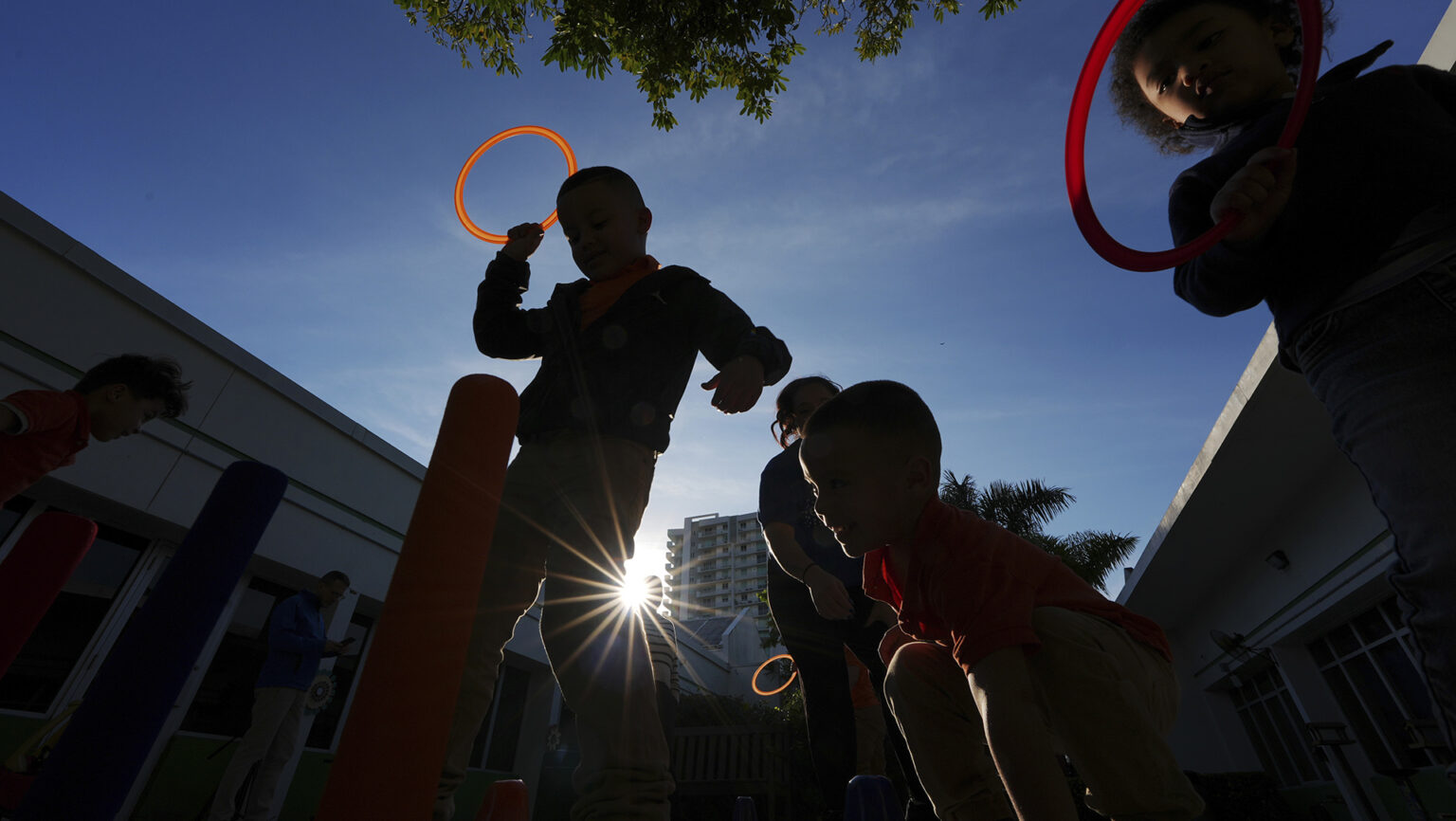 Multiple children playing with plastic ring toss toys are seen in silhouette from below, illuminated by sunrays in a courtyard surrounded by buildings and under foliage and a partly cloudy sky.