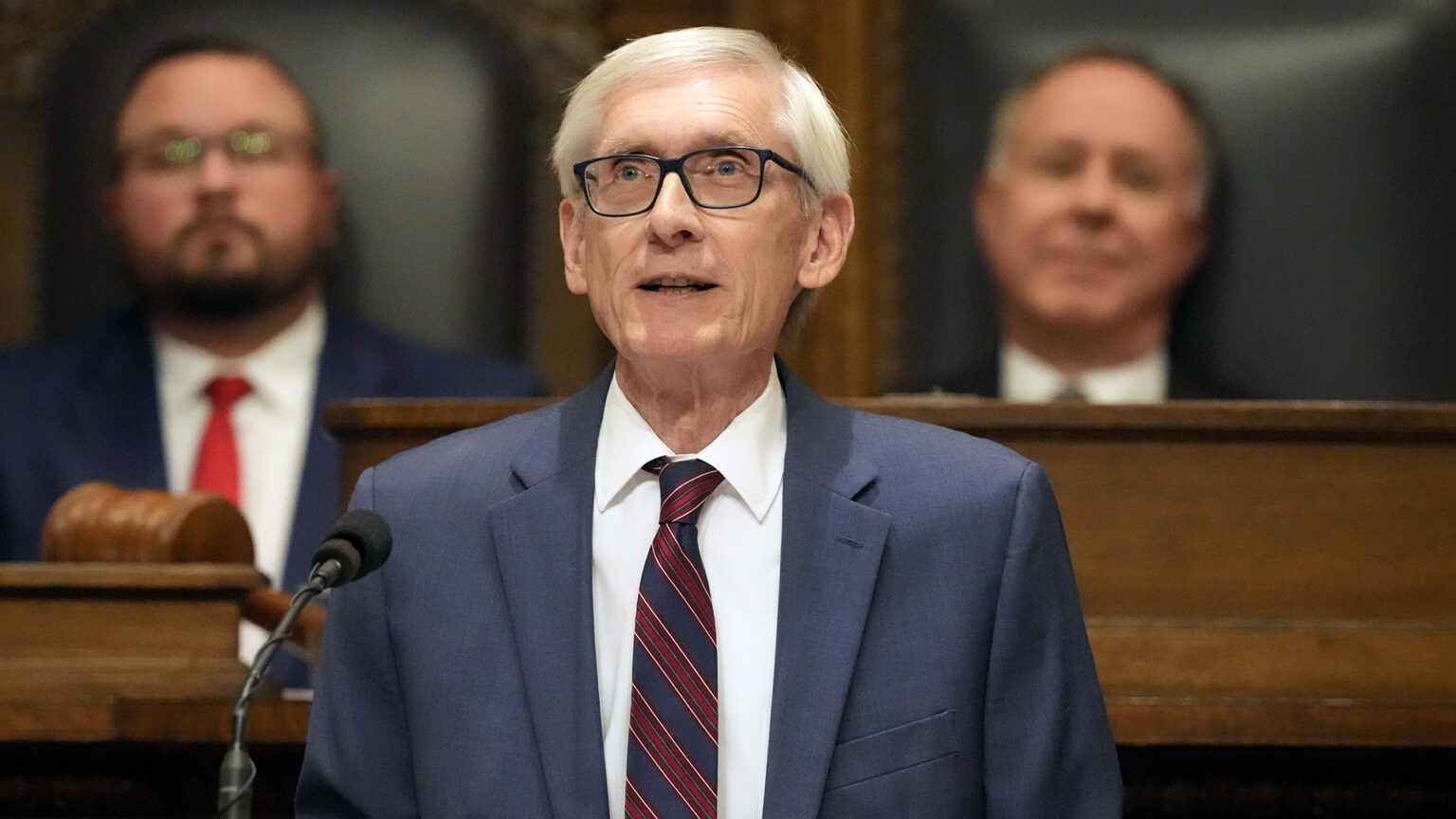 Tony Evers stands and speaks into a microphone, with two out-of-focus people seated in high-backed leather and wood chairs on a wood legislative dais in the background.