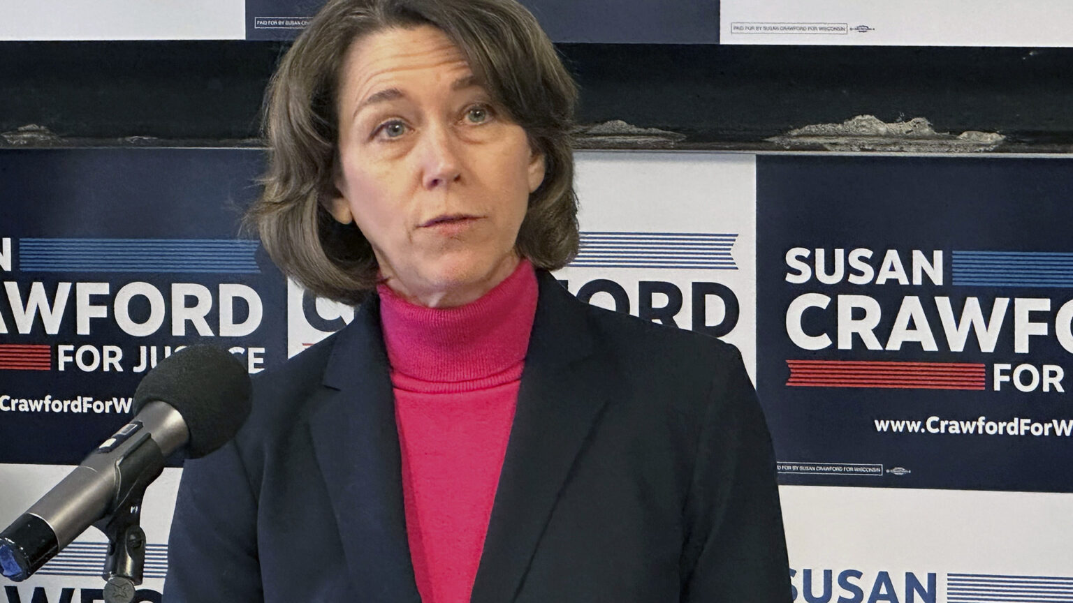 Susan Crawford speaks into a mounted microphone while standing in front of a backdrop consisting of multiple campaign signs in rows.