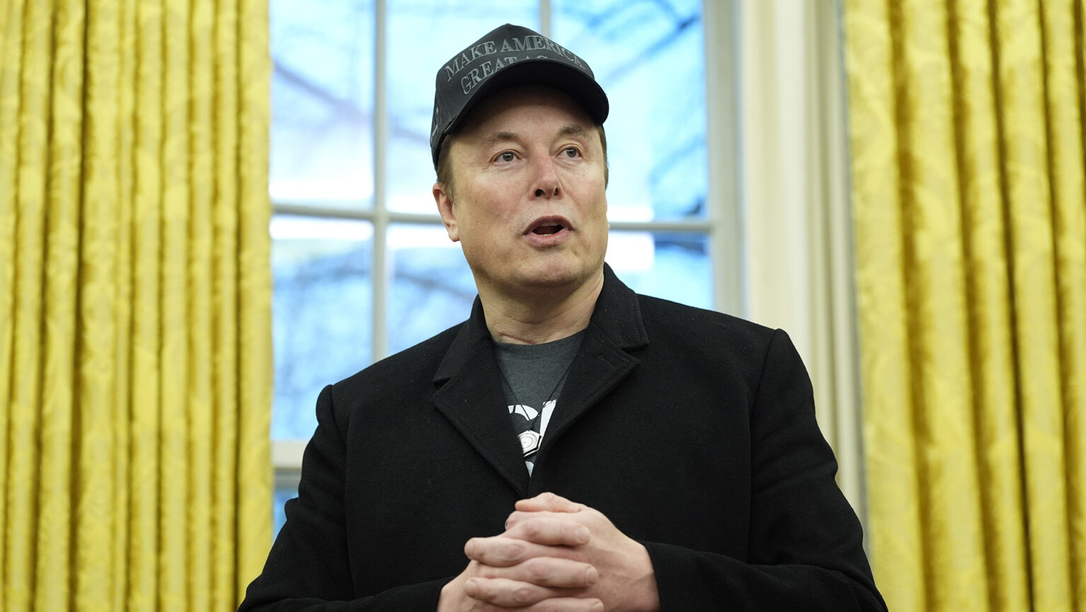 Elon Musk speaks with his hands folded in front of him while standing in front of a window with open curtains.