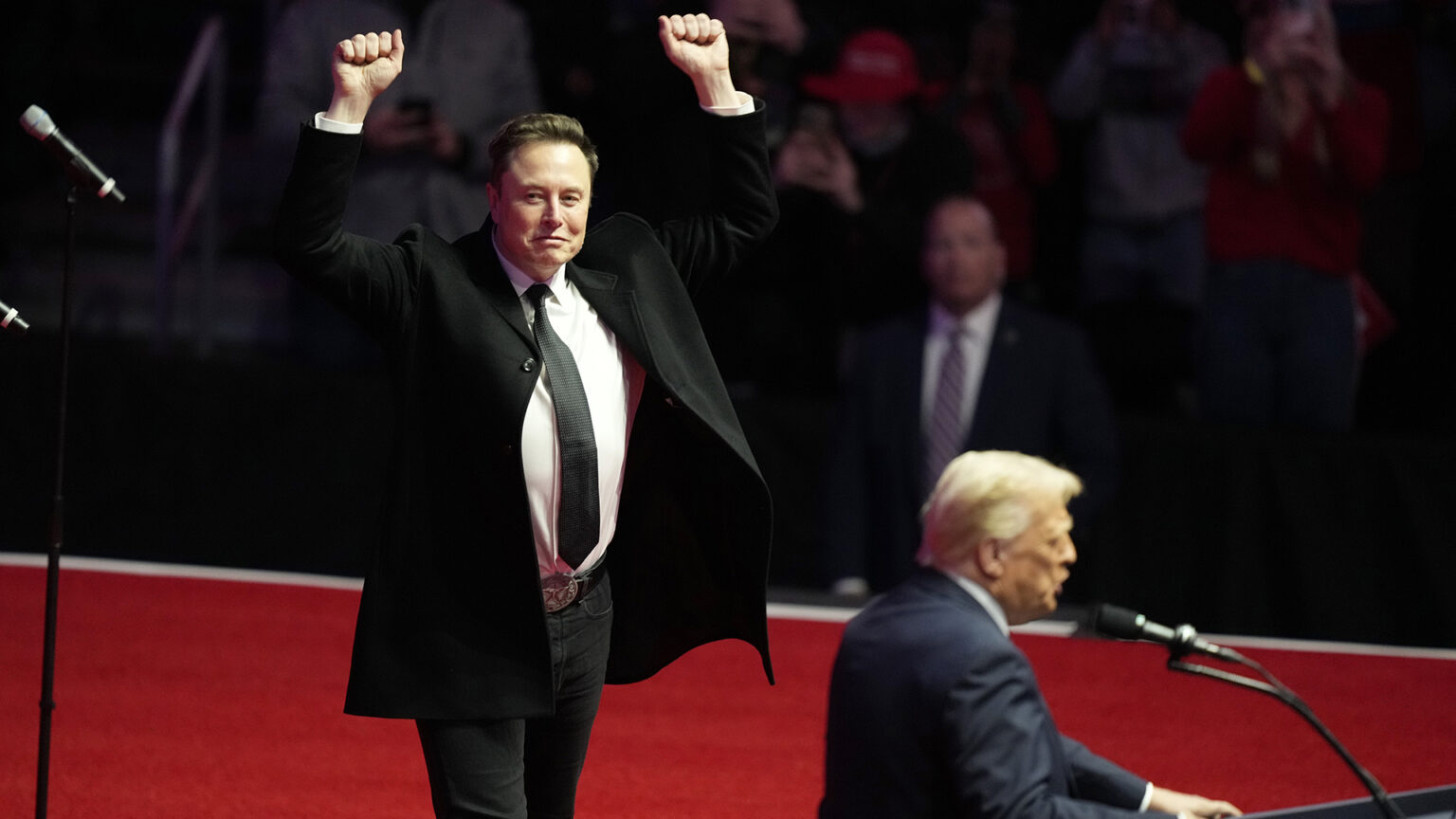 Elon Musk holds both of his arms up in the air and makes fists while standing behind an out-of-focus Donald Trump, who is speaking into a microphone mounted to a podium, with out-of-focus people seated and standing in the background.