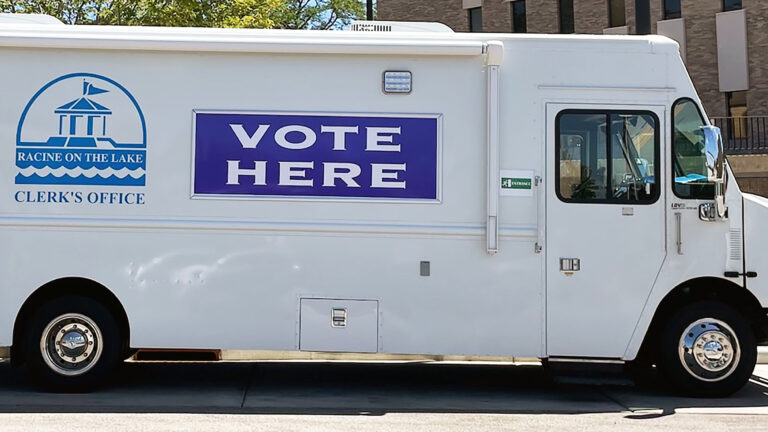 A step van with the logo of the City of Racine Clerk's office logo and the words VOTE HERE painted on its side is parked in a parking lot, with trees and a building in the background.