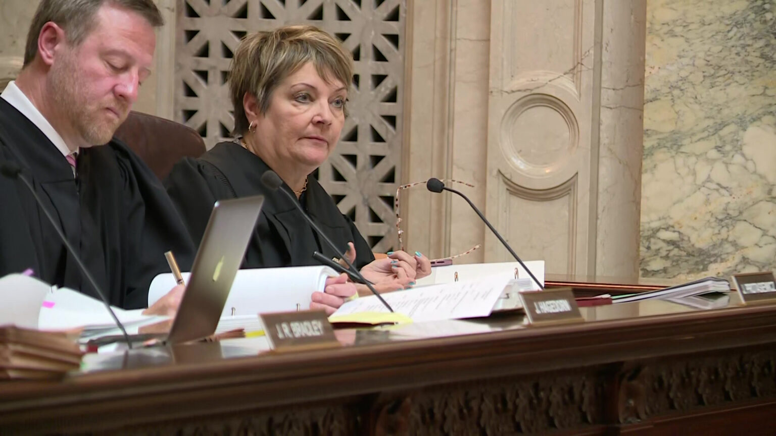 Janet Protasiewicz gestures with her right hand and holds eyeglasses with her right hand while seated in a high-backed leather chair and facing a microphone mounted to a wood judicial bench covered with papers, books, a laptop computer and other items, with Brian Hagedorn seated to her side in a room with marble masonry.