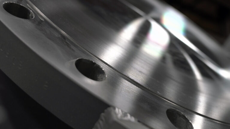 Light reflects off the surface of a multi-inch thick brushed stainless steel disk with drilled holes around its outer edge.