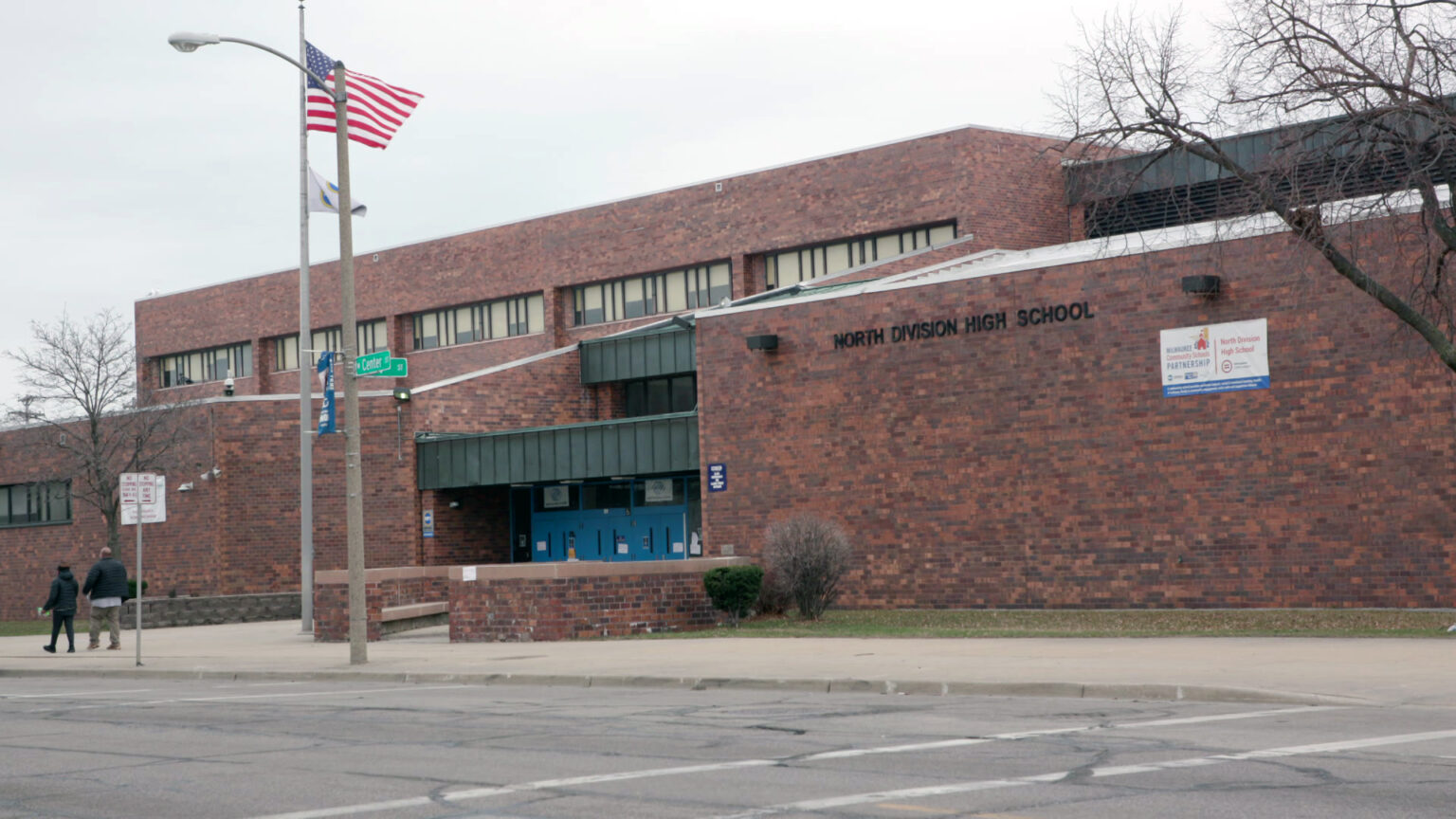 Two pedestrians walk along a sidewalk in front of a multi-story brick building with a letter sign reading North Division High School and an entrance with multiple double doors, with a street sign, flagpole with the U.S. flag and leafless trees in front of the structure.